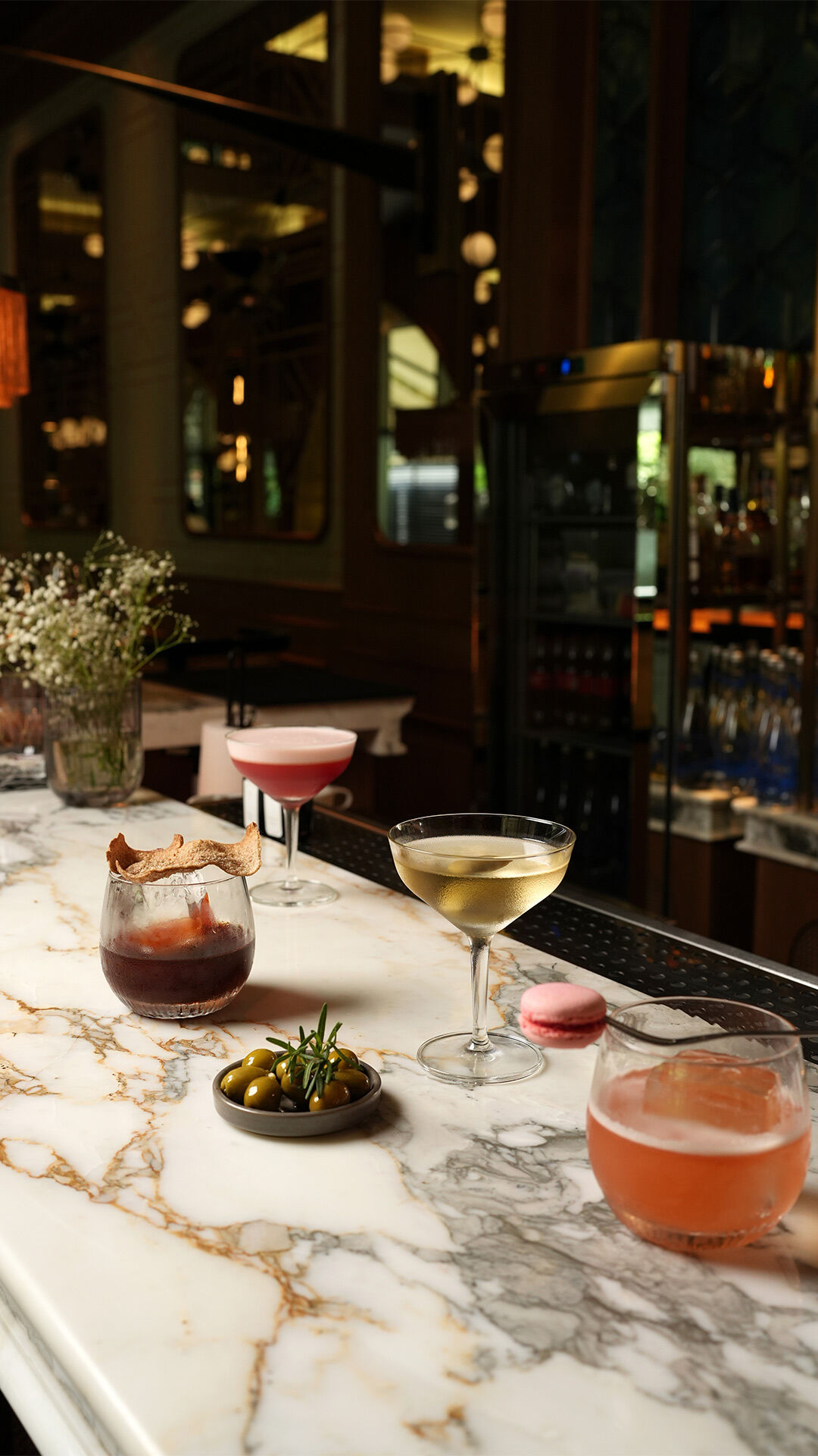 Elegant marble bar setup with assorted cocktails, a plate of olives, a pink macaron, and a vase of baby's breath flowers at a wedding reception.